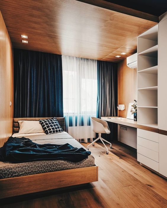 A bold minimalist teen room with a wooden ceiling with lights, navy curtains, a storage unit with a built in desk, a comfy bed and blue bedding