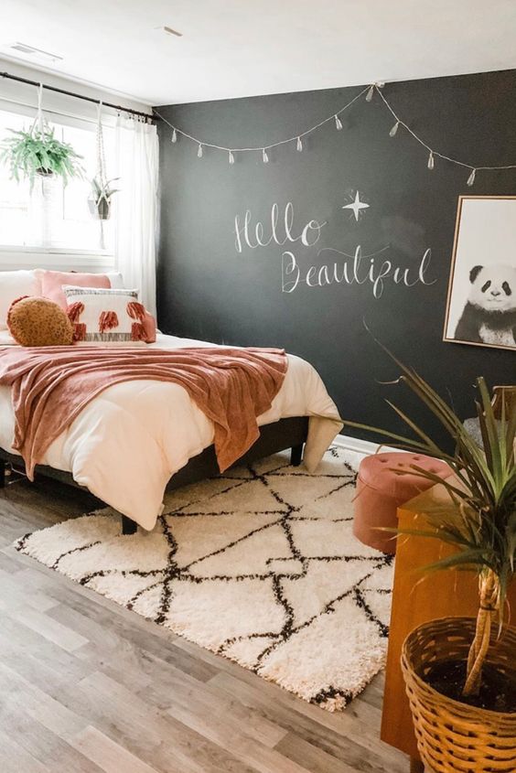 a chic teen bedroom with a chalkboard wall, a bed with pretty pink bedding, a wooden desk, a chair and a footrest