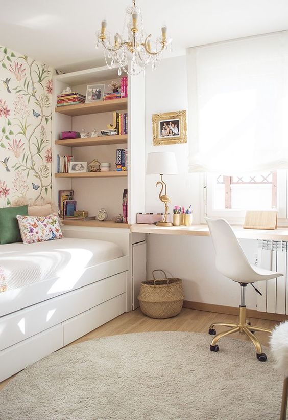 An airy teen room with floral wallpaper, built in shelves, a windowsill desk, a white chair and a crystal chandelier