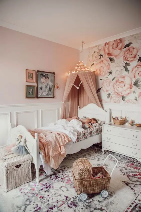 a beautiful pink girl's bedroom with floral wallpaper, white paneling, vintage white furniture, a printed rug and a dusty pink canopy over the bed