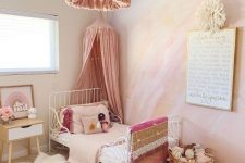 a dreamy pink girl’s bedroom with a watercolor yellow and hot pink accent wall, a white metal bed with pink bedding, a pink tassel chandelier