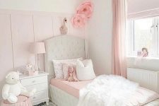 a light pink and white girl’s bedroom with a white upholstered bed with pink and bedding, a white nightstand, a pink pouf and a crystal chandelier