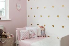 a pretty girl’s bedroom with a light pink wall, a gold heart accent wall, a white bed with pink and white bedding, a basket with toys