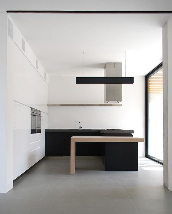 A minimalist black and white kitchen with a white storage unit and upper cabinets, black lower ones and a kitchen island with a light stained table