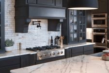 a modern black and white farmhouse kitchen with sleek matte cabinets, a large vintage hood, white marble countertops and a white marble tile backsplash, black lamps