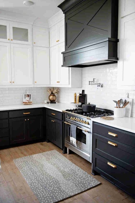 a modern farmhouse kitchen with black lower cabinets and white upper ones, a large hood covered with wood and gold fixtures