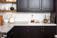 a rustic meets modern kitchen with black shaker cabinets, white stone countertops and a white tile backsplash, brass fixtures