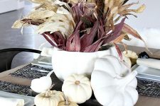 a Thanksgiving centerpiece of a white soup bowl with wheat, feathers, corn husks and some white pumpkins around it