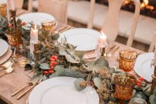 a chic Thanksgiving table with a greenery and berry runner, candles, amber glasses and woven chargers plus gold cutlery