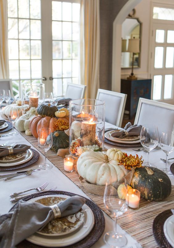 a chic rustic Thanksgiving table with a wooden runner, woven chargers, pritned plates, grey napkins and lots of pumpkins and blooms