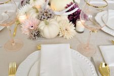 a glam Thanksgiving table setting with a centerpiece of white pumpkins and neutral and purple blooms, candles, gold cutlery and blush glasses