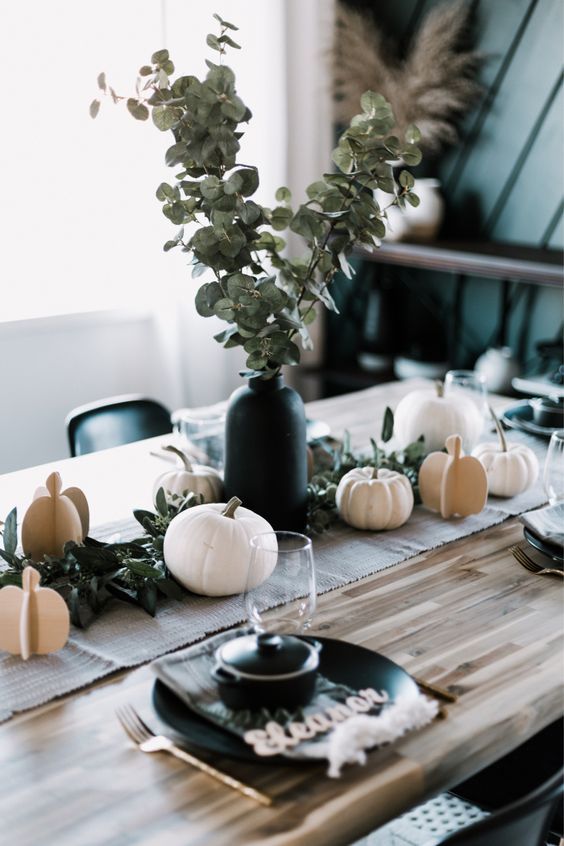 A modern Thanksgiving table with white pumpkins   real and cardboard ones, a greenery arrangement in a black vase and black plates