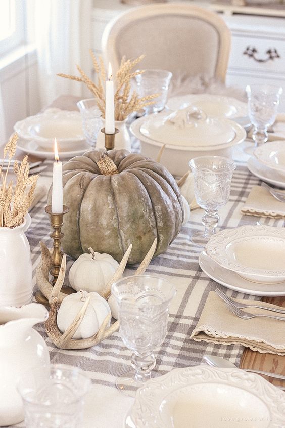 a neutral farmhouse Thanksgiving table with a plaid tablecloth, neutral pumpkins, antlers, candles and wheat in jugs is very beautiful
