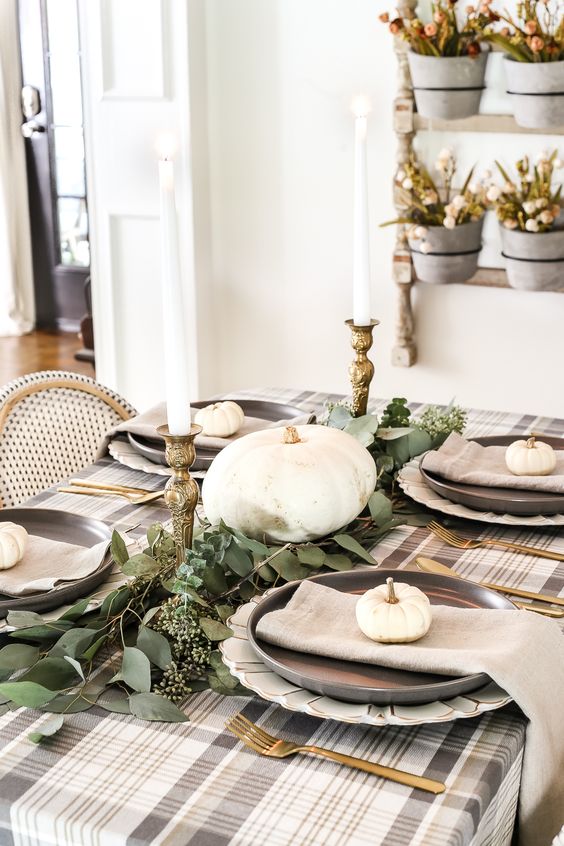A neutral low key Thanksgiving tablescape witha plaid tablecloth, grey napkins, a greenery runner, white pumpkins and candles
