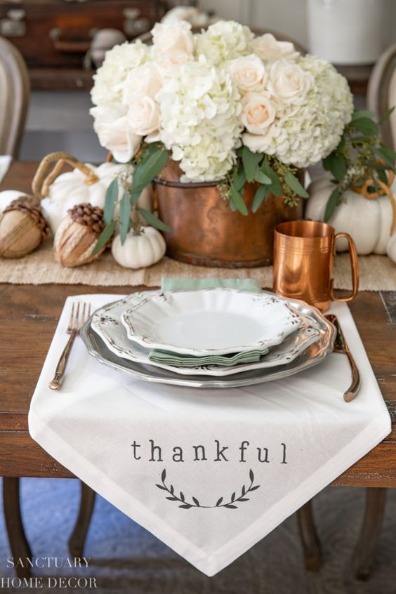a neutral rustic Thanksgiving table with a plaid runner, a white napkin, a copper mug, a bucket with hydrangeas and roses and acorns