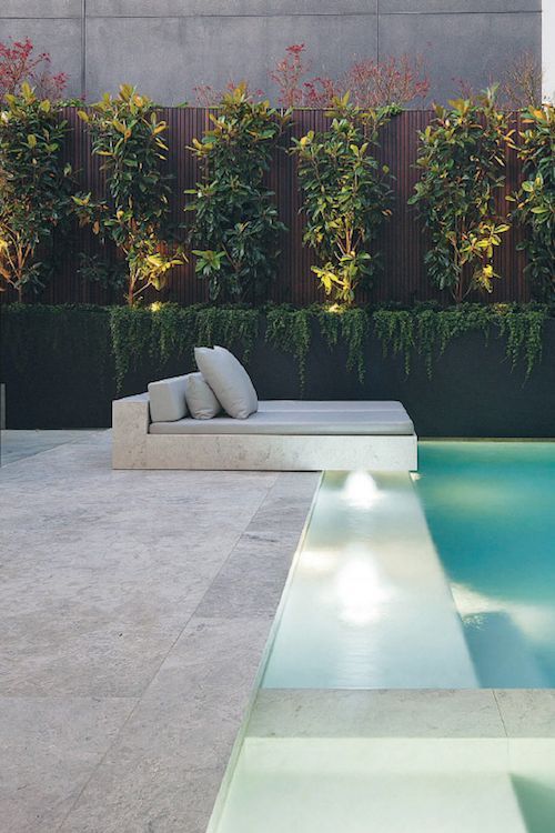 A minimalist outdoor space with a large lit up pool, a neutral stone deck and a built in bed with neutral bedding