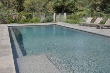 a neutral outdoor space with a blue pool, stone tiles and neutral loungers plus greenery around is amazing for spending time here