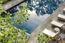 a soothing outdoor space with a pool, a stone deck and neutral loungers, a dining space and greenery around