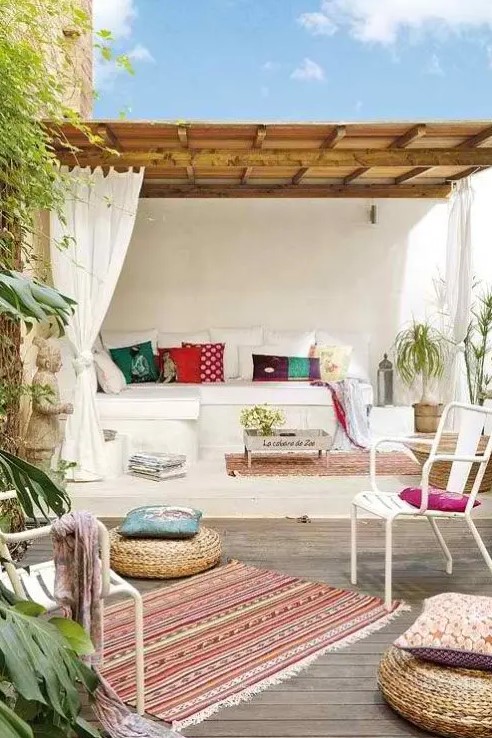 a beautiful and welcoming Mediterranean outdoor space with a white built-in sofa with bright pillows, a roof over this seat, colorful rugs and pillows and white chairs and jute poufs