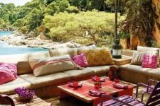 a bright and colorful outdoor living room with neutral upholstered benches, colorful pillows, bright stools and textiles plus a sea view