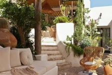 a chic Mediterranean outdoor living room done in neutrals, with a roof, greenery, a creamy sofa and pillows, stumps and a wicker chair, pendant lamps