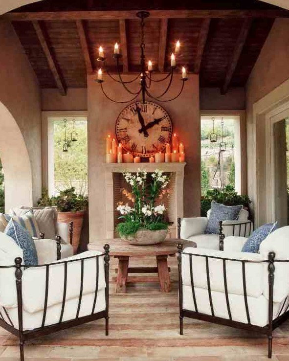 A cozy Mediterranean living room, outdoors and indoors, with a non working fireplace, a round wooden table, metal chairs with white upholstery and greenery