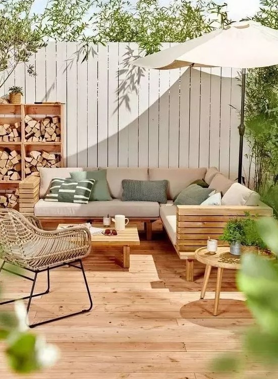 a lovely modern outdoor living room with a wooden deck, a planked sofa and a table, a rattan chair, a storage unit with firewood