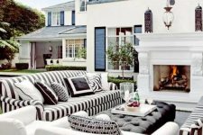 a lovely outdoor living room with a fireplace, striped black and white sofas, white chairs, a black upholstered ottoman and some greenery