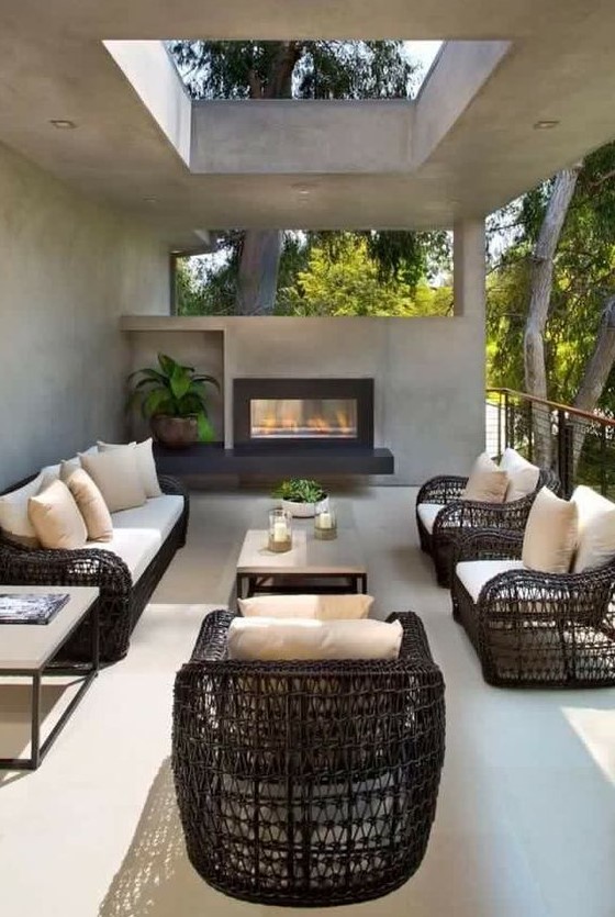 A modern outdoor living room with black wicker furniture, a concrete coffee table, a built in fireplace and some potted greenery and a skylight