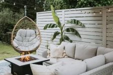 a small and cool farmhouse outdoor living room with a neutral wicker corner sofa, a fire pit, a pendant egg chair and a potted plant