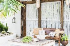 a welcoming tropical patio with woven lanterns, a wooden beach with pillows and a vintage low table plus cacti in a flower bed
