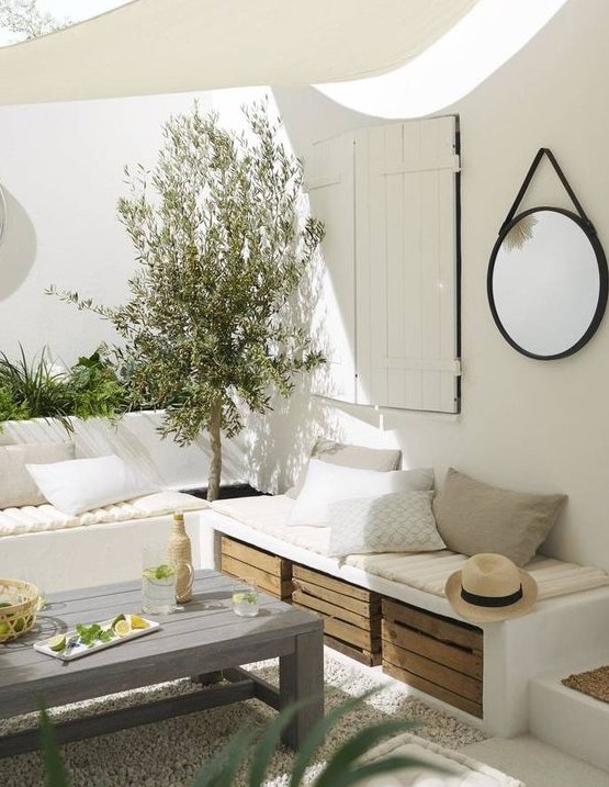 A white Mediterranean terrace with built in benches, greenery and a tree, crates for storage and a small roof