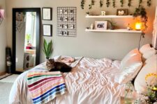 a neutral boho bedroom with greige walls, a bed with neutral bedding, open shelves, hanging greenery, a nightstand with blooms
