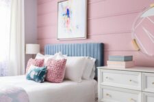 a stylish pastel teen bedroom with a pink panel accent wall, a blue upholstered bed, a white dresser, a mirror and a sunburst chandelier