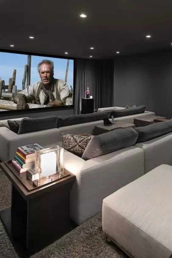 A modern home theater with graphite grey walls, neutral sofas and grey pillows, a large screen, built in lights and a side table