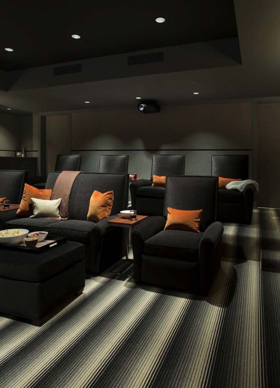 A moody home theater with graphite grey walls and a ceiling with built in lights, black chairs and sofas, bright pillows