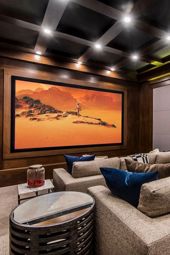 A stylish and chic home theater with stained wooden walls, grey seating furniture, a large screen, built in lights, shiny side tables