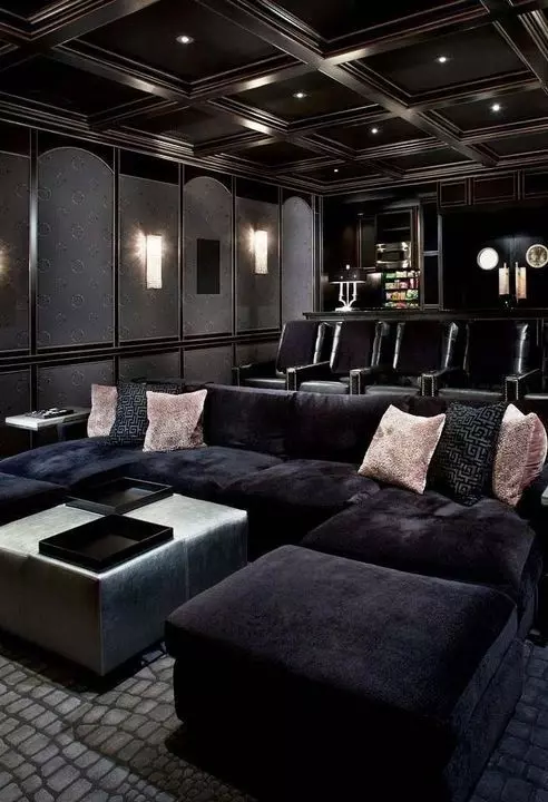 An elegant moody home theater in black, with a large sofa and lots of comfrotable chairs, a pouf and pink pillows, built in lights