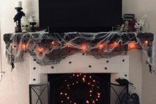 a black Halloween mantel with black and white spider web, lights, spiders, a lit up wreath and candleholders plus bats over the mantel