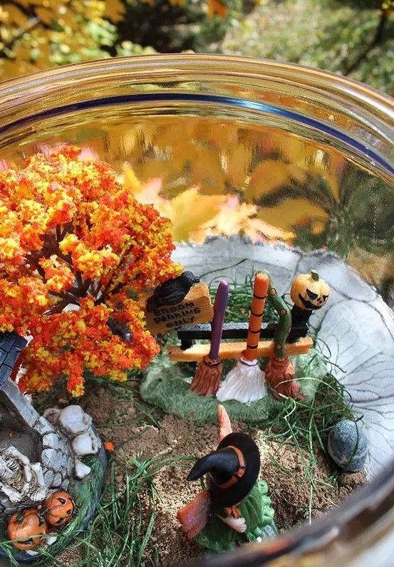 A fun Halloween terrarium with a witch, a fall tree, jac o lanterns, skulls and bones and brooms at a stand