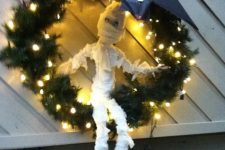 a funny Halloween mummy decoration on a Christmas wreath with lights and with a black bat on top to save it from rain