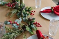 a bold Christmas table setting with an evergreen and pinecone runner, a pillar candle and red napkins plus berries