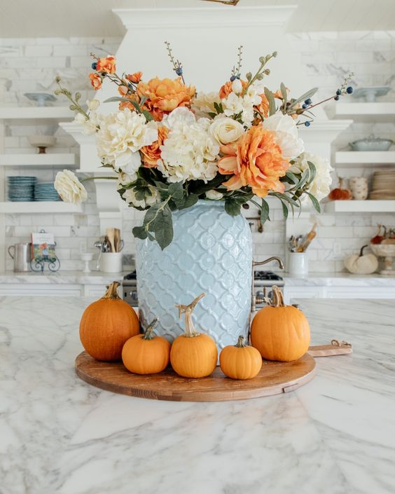 a bold modern Thanksgiving centerpiece of a blue jar, white and orange blooms and orange pumpkins around for Thanksgving