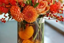a bright fall or Thanksgiving centerpiece of a clear vase with orange gourds, bold orange blooms and berries is a very fresh and vibrant idea