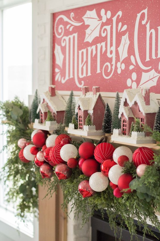 a catchy Christmas mantel decorated with evergreens, bold red and white ornaments, pink houses and bottle brush Christmas trees