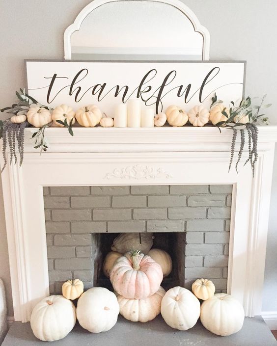 a chic white Thanksgiving mantel with pumpkins, long flowers, white candles, a sign and a mirror behind it