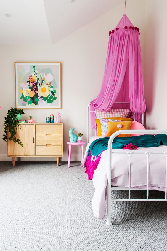 a colorful and welcoming kid's room with a bed with colorful bedding and a hot pink canopy, a dresser, a pink stool and some floral artwork