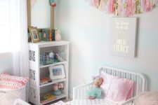 a pastel kid’s room with light green walls, a white bed with colorful bedding, a white storage unit and colorful tassels