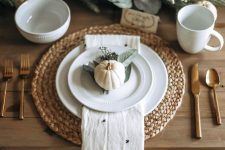 a pretty and chic Thanksgiving place setting with a woven placemat, white porcelain, a printed napkin, a pumpkin and some leaves is pure chic