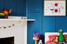 a quirky kid’s room with blue paneling on the walls, a white birkc fireplace used for book storage, a bed with colorful bedding and toys